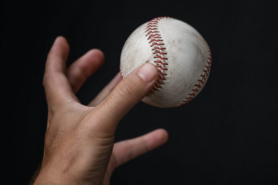 A hand catching a used white baseball with red stitching against a black background